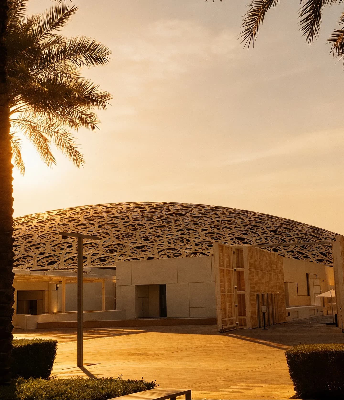 The dome of the Louvre Abu Dhabi at golden hour, seen from outside between two palms