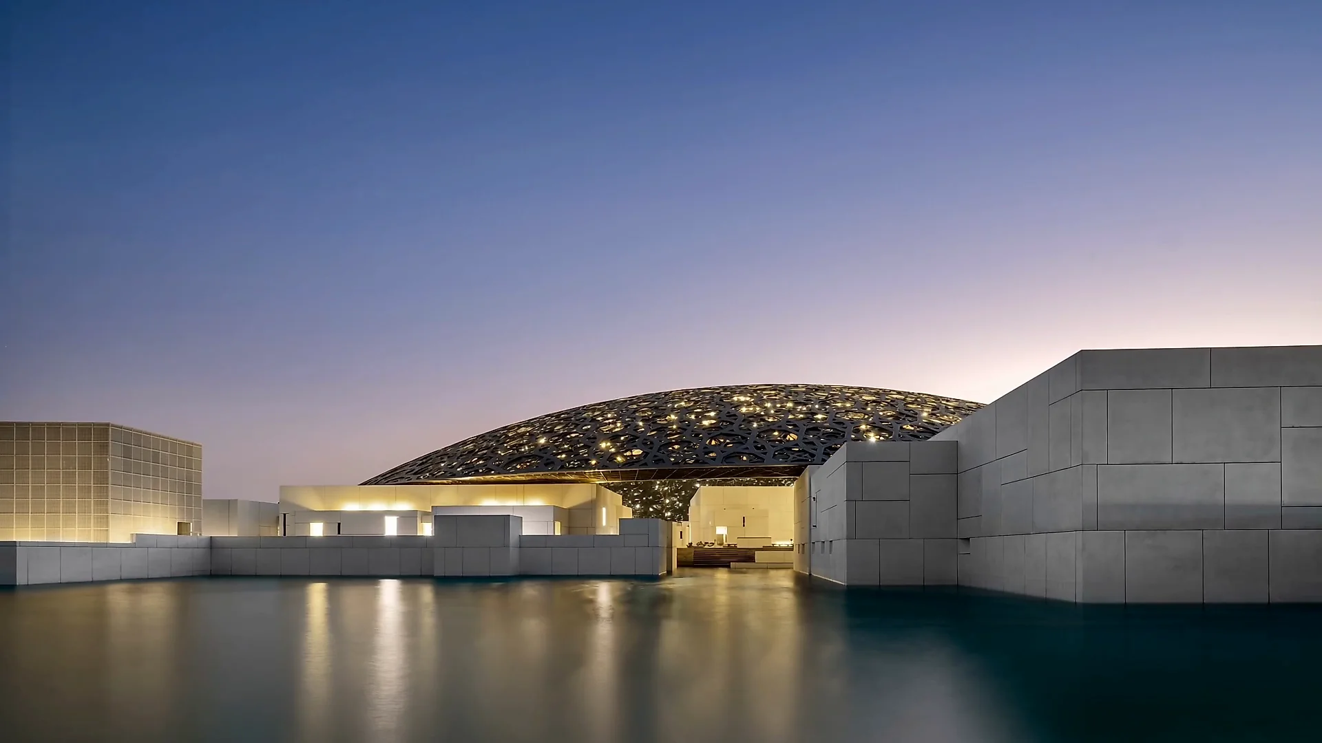 The Louvre Abu Dhabi at dusk, seen across the water.