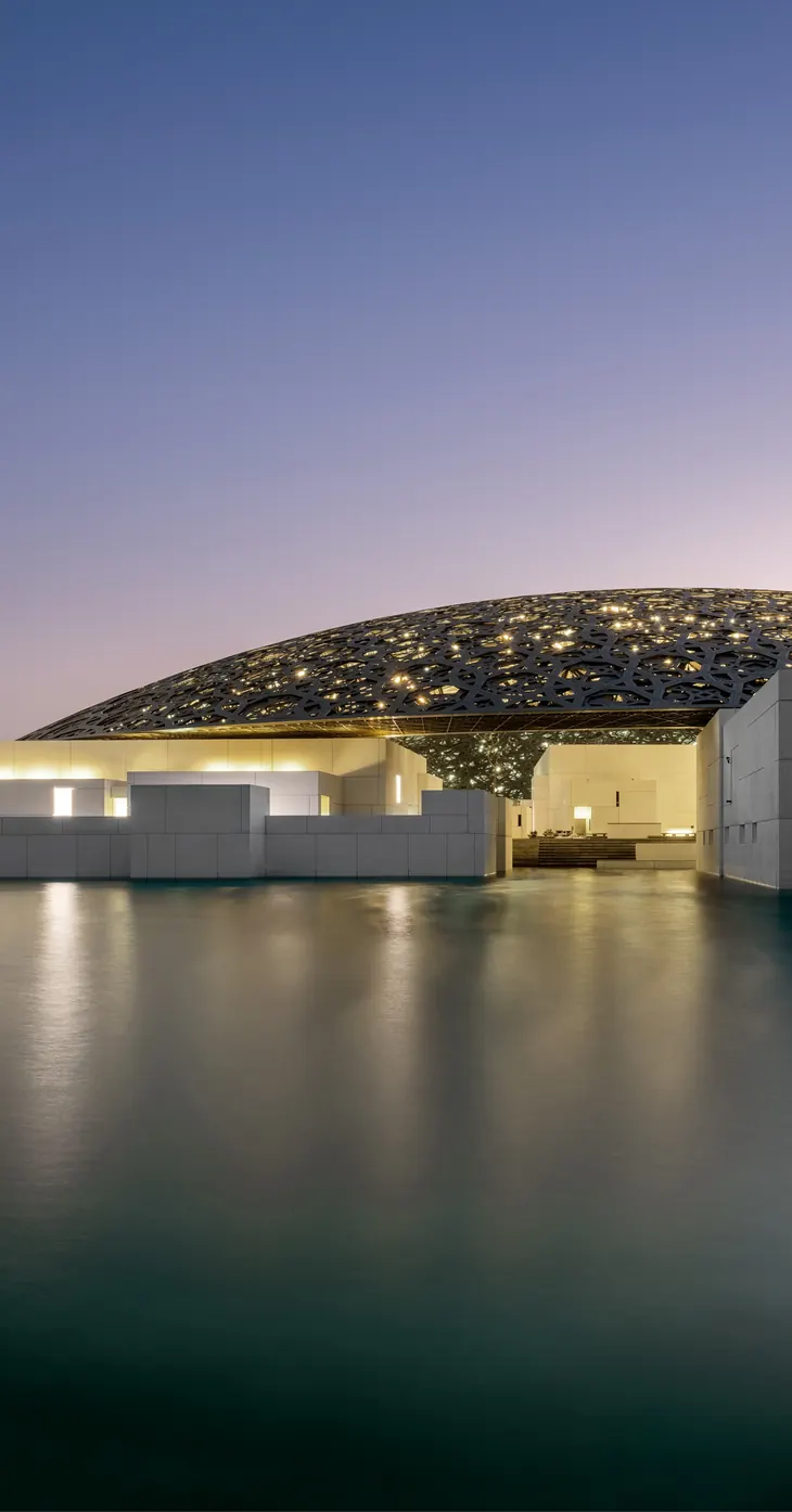 The Louvre Abu Dhabi at dusk — its latticed dome glowing low over still water, the lit galleries beneath it